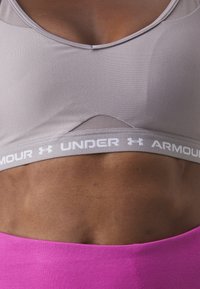 Grå sportbehå med nätpanel, med ett vitt "Under Armour"-logoband. Matchad med en ljusrosa högmidjad plagg som visar upp texturerad tyg.