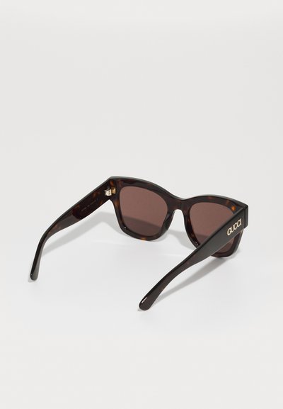  schildpadbril met bruine glazen, met een slank cat-eye ontwerp. Gouden logo detail op de veren. Gladde textuur en hoekige vorm.