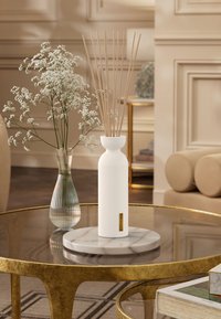 Vit ribbad keramisk diffuser med bambustickor på en marmorbass, placerad på ett glasbord med guldaccenter och en blomsterarrangemang.