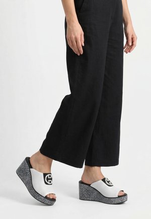 Person trägt weiße und schwarze Slip-On Keilsandalen mit gemusterten Sohlen und schwarze Hosen mit weitem Bein, stehend vor weißem Hintergrund.
