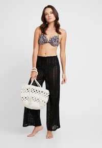 Μπικίνι με λεοπάρ μοτίβο, διάφανα μαύρα κροσέ παντελόνια και λευκή πλεκτή τσάντα με περίπλοκες λεπτομέρειες. Μοντέλο στέκεται ξυπόλητο σε απλό φόντο.
