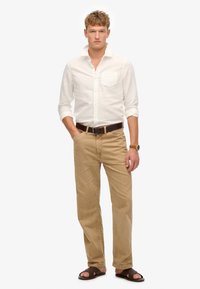 Camisa blanca de manga larga, chinos beige, cinturón marrón y sandalias marrón oscuro. La camisa tiene un bolsillo; los chinos están ligeramente desgastados.