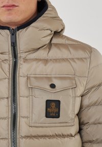 Giubbotto puffer beige con cappuccio, design trapuntato, zip frontale e taschino sul petto con una patch logo nera e arancione. Texture liscia.