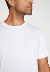 Fehér sport póló sima, könnyű anyagból. Kerek nyakkivágással és rövid ujjakkal rendelkezik, a mellkasán pedig egy diszkrét szürke logó található.