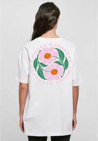 Hvit T-skjorte med et stort trykk på ryggen som viser rosa blomster, grønne blader og teksten "Velg vennlighet" i rosa og svart.