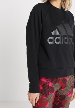 Vrouw draagt een zwarte Adidas-sweatshirt en legging met geometrisch patroon in rood, roze en bruin, staand tegen een effen lichte achtergrond.
