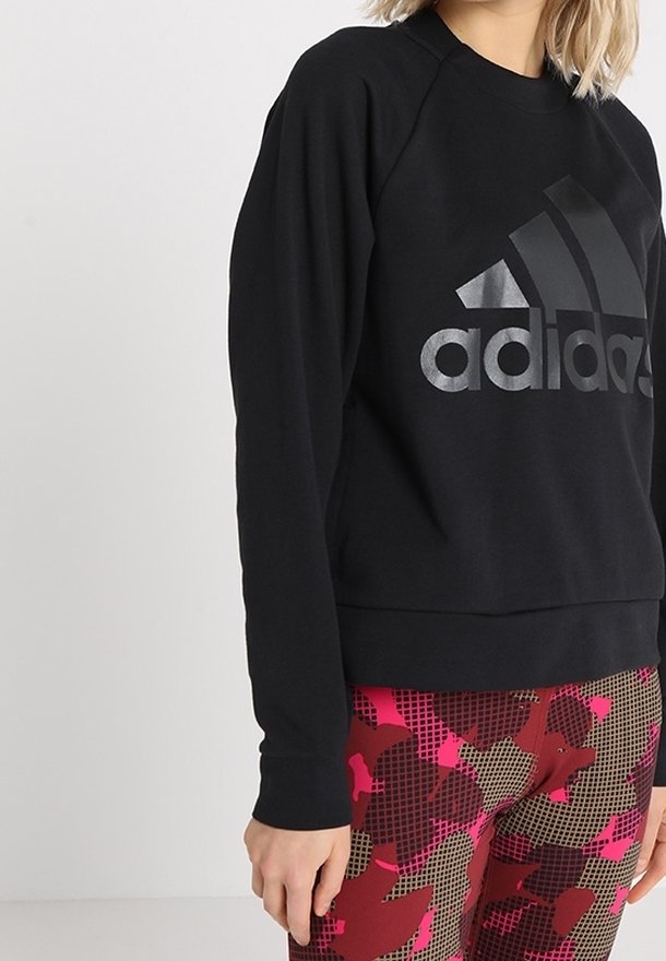Femme portant un sweat-shirt noir Adidas et des leggings à motifs géométriques rouges, roses et marron, debout devant un fond clair uni.