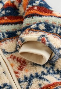 Détail d'une veste en polaire présentant un motif coloré et texturé dans des teintes de bleu, orange et crème, avec un poignet doux et roulé.