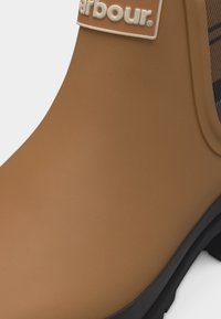 Κοντινό πλάνο μπεζ αδιάβροχου μποτάκι με μαύρη σόλα και υφασμάτινο πάνελ που δείχνει μοτίβο τάρταν και λογότυπο της μάρκας.