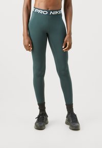 Zelene tajice koje se prilagođavaju tijelu s crnim elastičnim pojasom na kojem piše "NIKE PRO" u bijeloj boji. Materijal je gladak, s elegantnim i aerodinamičnim dizajnom.