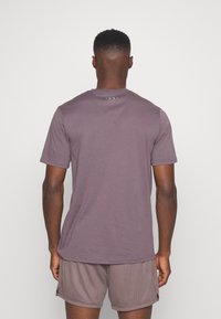 Hombre con pantalones cortos deportivos morados y camiseta de manga corta a juego, de pie frente a un fondo blanco con la espalda visible.