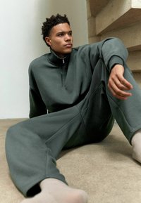 Sudadera de cuello con cremallera en verde oscuro y pantalones de pierna ancha a juego, hechos de una tela suave, con puños acanalados y un ajuste relajado.