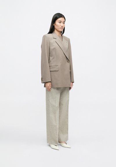 Υπερμεγέθη μπεζ σακάκι με πέτο και μπροστινές τσέπες, σε συνδυασμό με ανοιχτό γκρι παντελόνι με φαρδιά πόδια και λευκά παπούτσια με μυτερή μύτη.