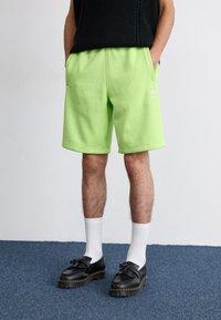 Shorts en coton vert néon avec une taille élastique, des poches latérales et un détail de logo blanc. Portés avec des chaussettes blanches et des chaussures noires.