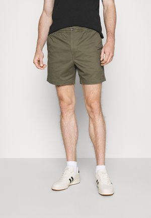 Modèle masculin portant un short vert olive, un t-shirt noir, des chaussettes blanches et des baskets blanches avec des rayures noires, debout devant un fond uni.