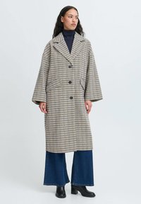 Langternet ternet frakke i beige, sort og blå, med dobbeltradet front, oversized krave og store lommer. Kombinationen er med vide jeans og støvler med hæl.