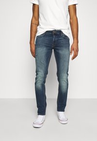 Jeans de ganga em azul escuro com área da coxa desbotada, ligeira desgastação na parte da frente e design de perna reta, combinados com ténis brancos.