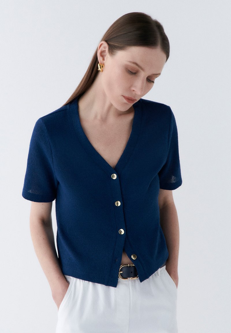 Marinblå kortärmad cardigan med V-ringning och guldfärgad knäppning, tillverkad av lätt material, med avslappnad passform och lätt textur.