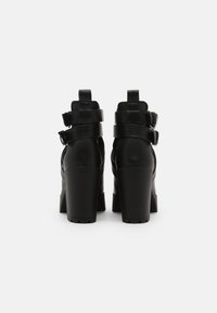 Zapatos de tacón alto negros con tacones gruesos, que cuentan con un diseño elegante, material texturizado y correas ajustables en los tobillos.