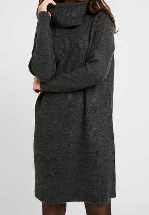 Woman wearing a dark gray ribbed knit turtleneck sweater dress with long sleeves, posing with one arm across her body.