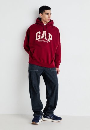 Hombre con sudadera con capucha roja de GAP Times Square, pantalones anchos de rayas finas y zapatillas deportivas azul marino, de pie contra una pared blanca mirando hacia la derecha.