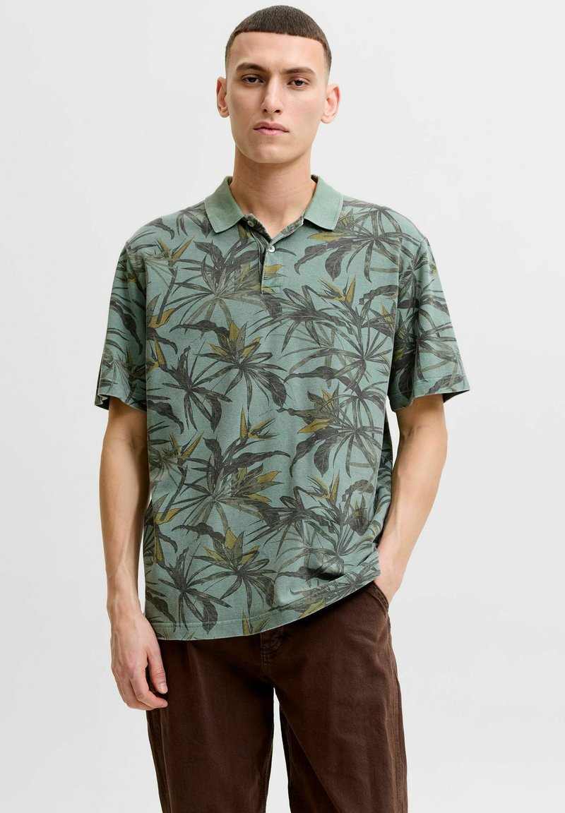 Jeune homme aux cheveux courts portant un polo vert à motif floral et un pantalon marron, debout avec une main dans la poche.