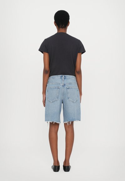 Shorts en denim bleu clair avec un ourlet effiloché et un design à cinq poches, associés à un T-shirt noir à manches courtes et des chaussures noires à enfiler.