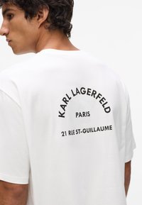 Biela bavlnená tričko s okrúhlym výstrihom. Text na zadnej strane obsahuje "KARL LAGERFELD," "PARÍŽ," a "21 RUE ST-GUILLAUME" v tučnom čiernom písme.