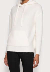 Felpa leggera in misto cotone beige con tasca frontale, cappuccio con cordino, texture a coste e spacchi laterali. Abbinata a pantaloni neri.