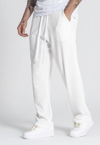 Pantalones de chándal blancos con textura, cinturilla elástica y cordón, con bolsillos laterales y un sutil detalle de logo. Combinados con zapatillas blancas.