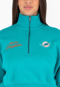 Felpa a mezza zip dei Miami Dolphins in un vivace teal. Presenta il nome della squadra ricamato in arancione e il logo con dettagli a raggi di sole. Materiale in morbido mix di cotone.