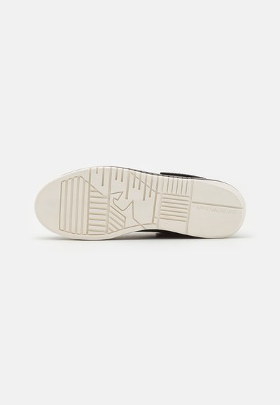 Black sneaker with a textured rubber sole featuring geometric patterns and an off-white base. Velcro strap at the top.