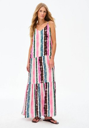 Woman with long blonde hair wearing a sleeveless, multicolored striped maxi dress and black sandals, standing against a white background.