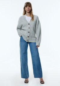 Grijze wollen cardigan met een iets oversized pasvorm, voorzien van vijf bruine knopen. Draag het met high-waisted blauwe jeans en zwarte hakken.