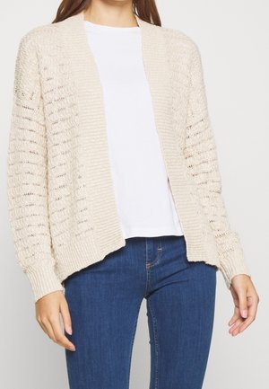Beżowy sweter typu kardigan z otwartym przodem, ribbowymi mankietami i teksturowanymi wzorami. Nosi się go na białą koszulę, w połączeniu z ciemnymi dżinsami.