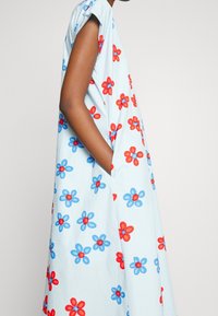 Personne portant une robe ample bleu clair avec des motifs floraux rouges et bleus, debout avec une main dans la poche latérale sur un fond uni.
