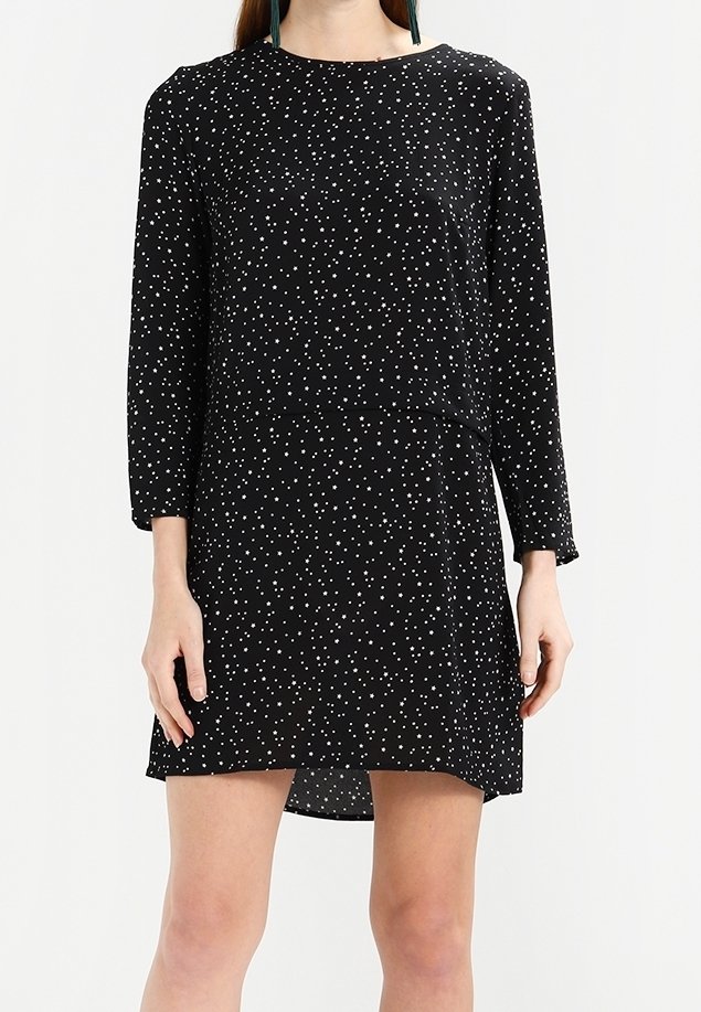 Femme portant une mini-robe noire à manches longues avec de petits pois blancs dispersés, debout devant un fond uni.