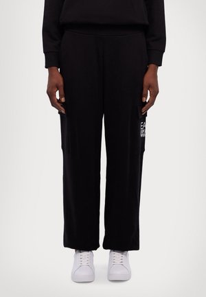 Person trägt schwarze weite Hosen mit Cargotaschen und weiße Turnschuhe, steht vor einem schlichten hellen Hintergrund.