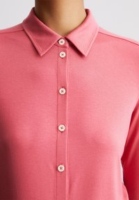 Rosa polo skjorte med spiss krage, med knappestolpe foran og lyse knapper, laget av myk, gl glatt stoff.