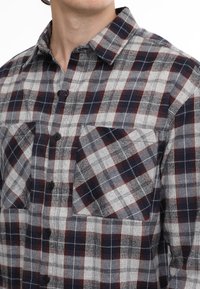 Camicia flanella a quadri rossa, blu navy e bianca con bottoni neri, colletto e due tasche sul petto. Texture morbida e vestibilità rilassata.