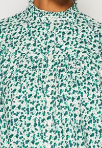 Weiße Bluse mit grünem und marineblauem Blumenmuster. Hat einen hohen, gerafften Kragen und eine durchgehende Knopfleiste mit kleinen weißen Knöpfen.