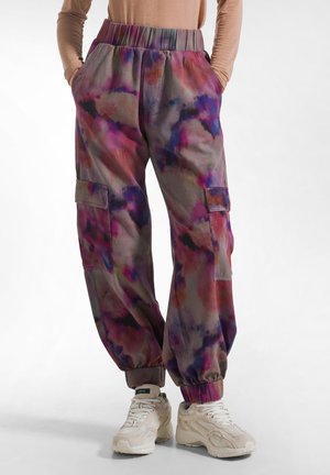 Person trägt pink-violette Batik-Cargohosen mit elastischen Bündchen und beigen Sneakers, steht mit den Händen in den Taschen.