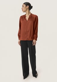 Blusa color ruggine con scollatura a V, spalle arricciate e maniche lunghe, abbinata a pantaloni neri a gamba larga e scarpe a punta.