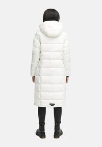Piumino lungo bianco con cappuccio, texture trapuntata, con etichetta del marchio all'orlo e pantaloni neri con stivali con lacci.