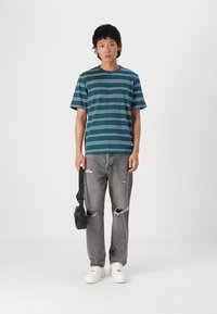 Camiseta de manga corta a rayas en tonos verde azulado y blanco, combinada con jeans grises rotos y zapatillas blancas, llevando un pequeño bolso negro.
