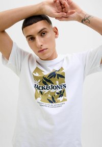 Hvid T-shirt med korte ærmer, der har et farverigt grafisk design med teksten "JACK & JONES" samt kamuflage-lignende bladmønstre i grøn og marineblå.