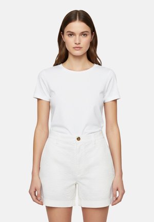 Donna con capelli castano lunghi che indossa una maglietta bianca a maniche corte semplice e pantaloncini bianchi texturizzati con un bottone marrone, rivolta in avanti.