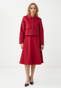 Czerwona bawełniana kurtka typu cropped z dwoma przednimi kieszeniami i guzikami, zestawiona z pasującą spódnicą w kształcie litery A. Modelka nosi czarne czółenka na szpilkach.