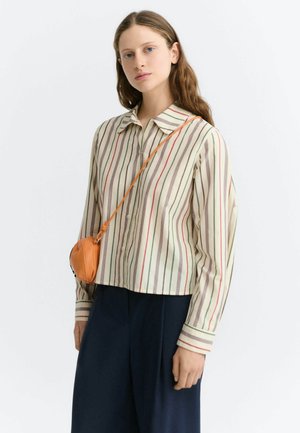 Jeune femme à la peau claire et aux cheveux bruns porte une chemise crème à rayures verticales multicolores et un pantalon large foncé, portant un petit sac bandoulière orange.