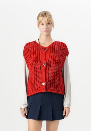 Vrouw in een chunky rode gebreide mouwloze cardigan met grote knopen over een beige blouse met lange mouwen en een marineblauwe geplooide rok.